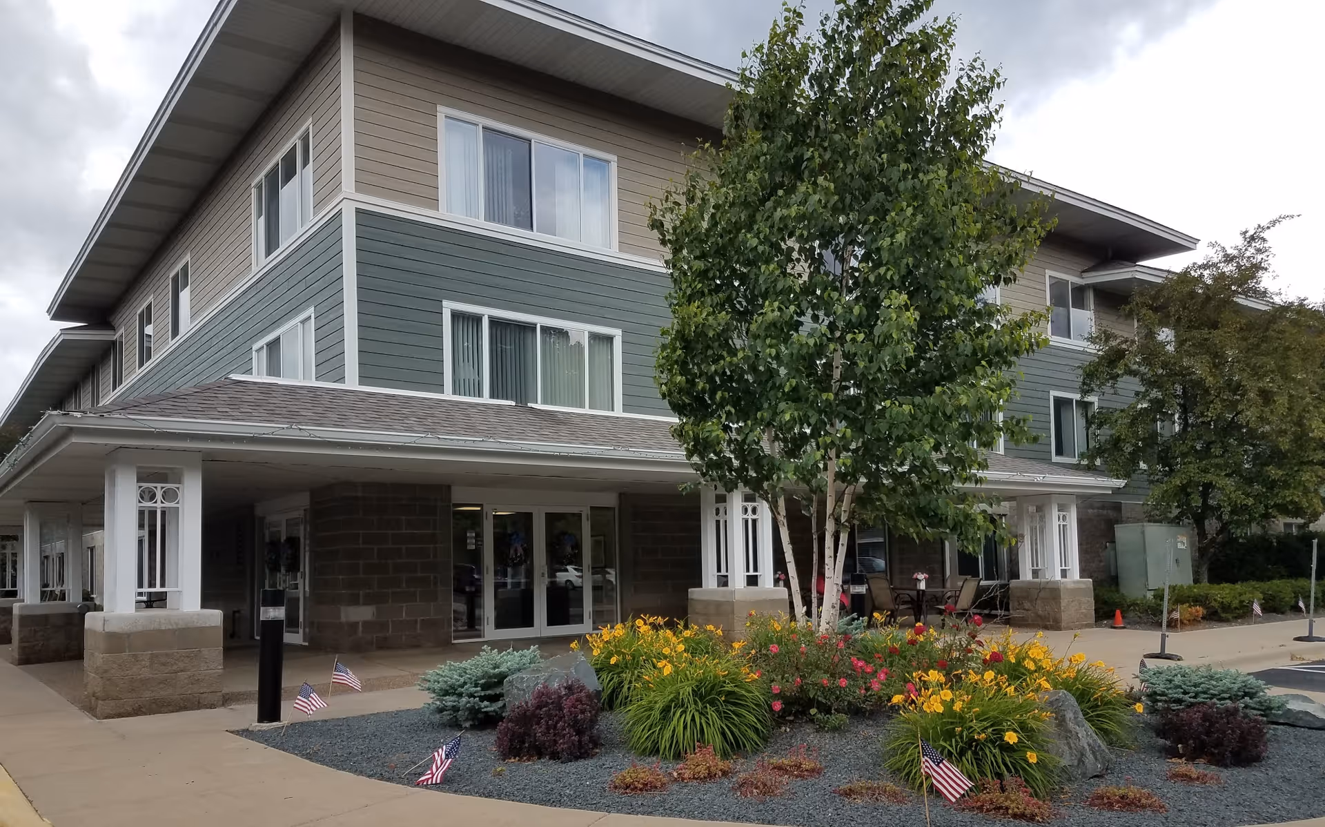 Exterior view of a multi-story senior living facility building with beige and green siding, large windows, and a covered entrance. In front of the entrance is a landscaped area with a variety of plants, flowers, small trees, and several small American flags. The sky is cloudy.