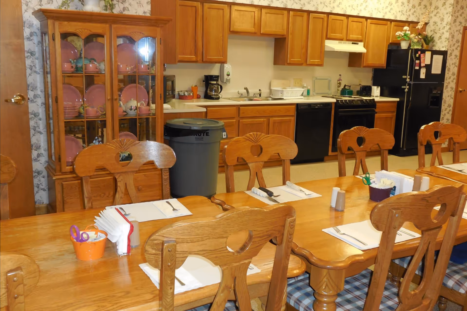 A dining area in a nursing home with a long wooden table set with placemats, napkins, forks, and knives. Wooden chairs with carved backs surround the table. In the background, there is a kitchen area with wooden cabinets, a black refrigerator, a stove, a dishwasher, a coffee maker, and a sink. A glass-front cabinet displays pink dishware and teapots. The walls have floral wallpaper.