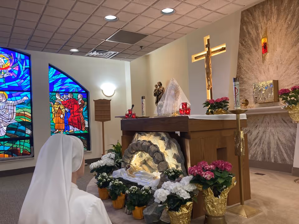 A nun dressed in white is kneeling in prayer inside a chapel. The chapel features a wooden altar adorned with flowers, candles, and religious statues. Behind the altar is a large crucifix mounted on the wall and a stained glass window depicting biblical scenes.