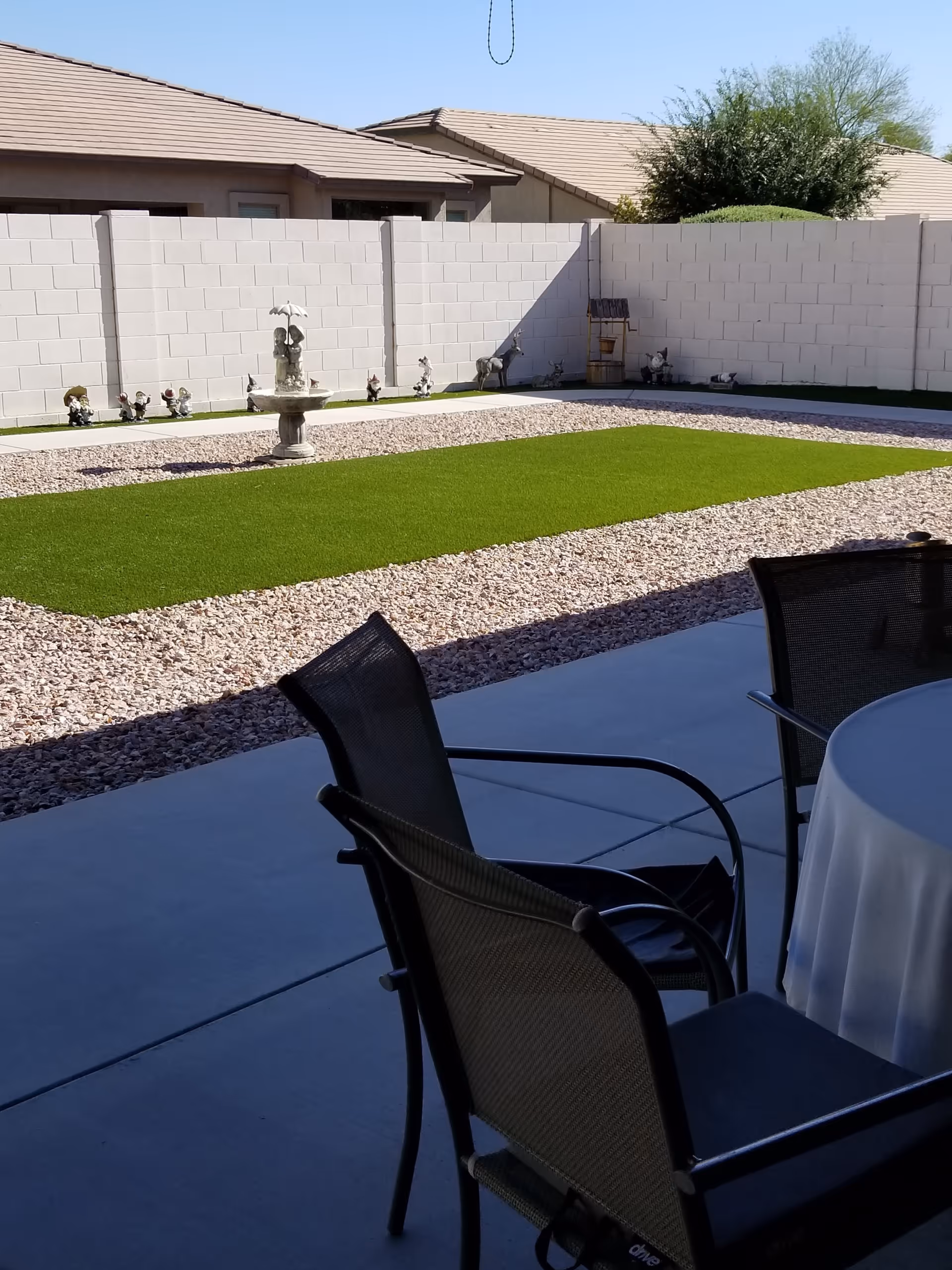 Outdoor patio area with a concrete floor, two black mesh chairs, and a round table covered with a white tablecloth. Beyond the patio is a landscaped yard with a rectangular patch of green artificial grass surrounded by small rocks. A white block wall encloses the yard, and there are various garden statues and a small fountain along the wall. The roofs of neighboring houses and some trees are visible in the background under a clear blue sky.