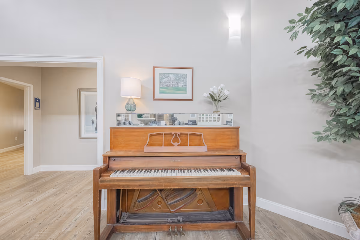 An upright wooden piano with a decorative music stand, placed against a light-colored wall. On top of the piano is a table lamp with a white shade and a glass base, a small rectangular mirror, a framed picture hanging on the wall, and a vase with white flowers. To the right of the piano is a large green leafy plant. The floor is light wood, and there is an open doorway to the left leading to another room.