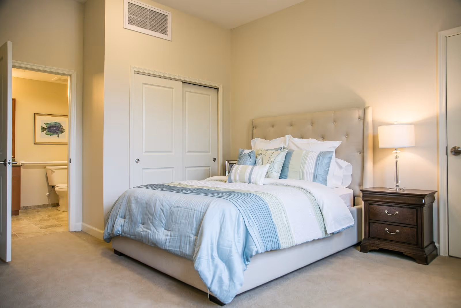 A neatly made bed with a tufted beige headboard and multiple pillows in shades of blue and white in a bedroom. To the right of the bed is a wooden nightstand with two drawers and a lamp with a white shade. To the left, an open door reveals a bathroom with a toilet and a framed fish picture on the wall.