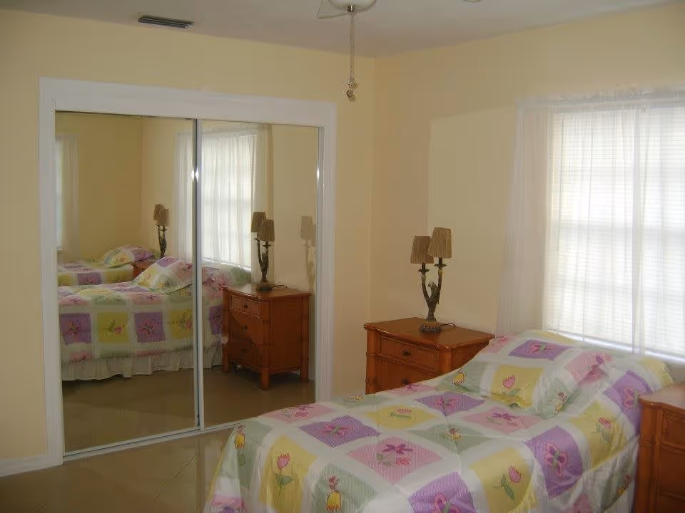 A bedroom with a single bed covered in a colorful quilt with floral patterns. There are two wooden nightstands on either side of the bed, each with a lamp. A large window with sheer white curtains lets in natural light. A closet with sliding mirrored doors is visible on the left side of the room.