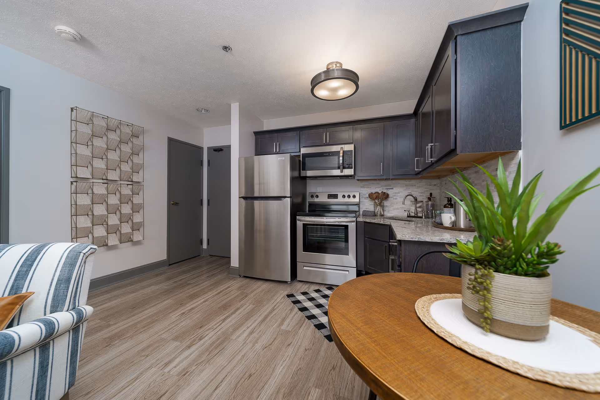 Modern kitchen area with stainless steel refrigerator, oven, and microwave. Dark wood cabinets and light granite countertops. A round wooden table with a potted green plant is in the foreground. Light wood flooring and a striped armchair partially visible on the left. Two closed gray doors and decorative wall art on the left wall.