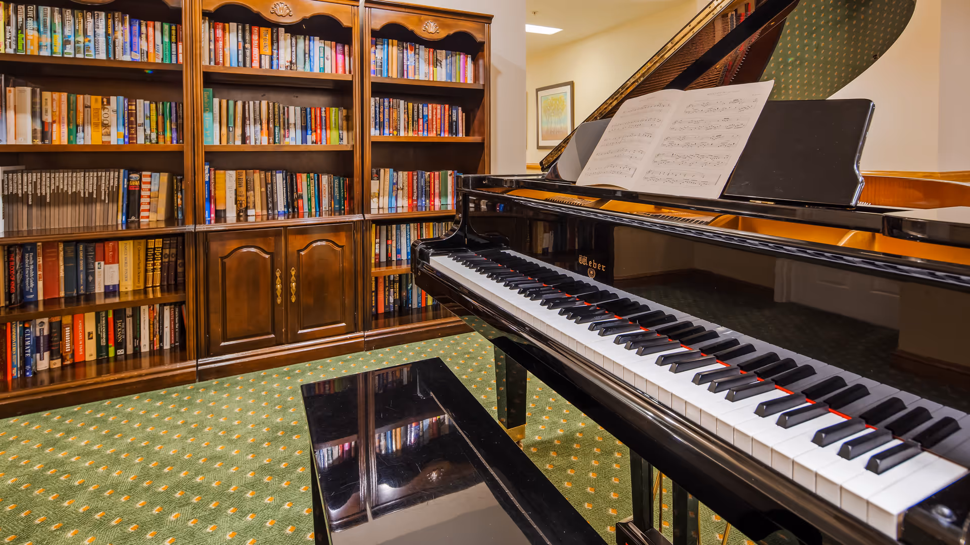 Interior view of a room with a black grand piano and an open sheet of music on the stand. Behind the piano, there is a large wooden bookshelf filled with books. The floor is carpeted with a green patterned carpet.