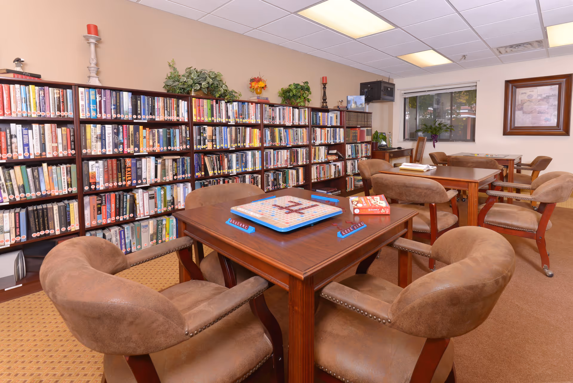 A cozy senior living facility library and game room with bookshelves filled with books along the wall. Several wooden tables with brown cushioned chairs are arranged in the room, with a Scrabble game set up on the nearest table. The room has beige walls, a window with a view outside, and decorative plants and framed artwork on the walls.