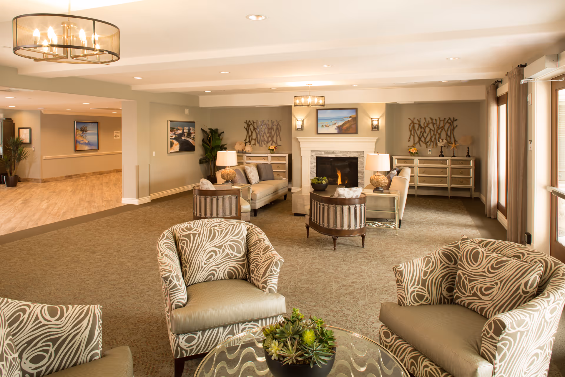 A spacious and well-lit living room area in a senior living facility featuring patterned armchairs around a glass coffee table with a plant centerpiece. In the background, there are sofas, side tables with lamps, a fireplace with a painting above it, and decorative wall art. Large windows with curtains allow natural light to fill the room.