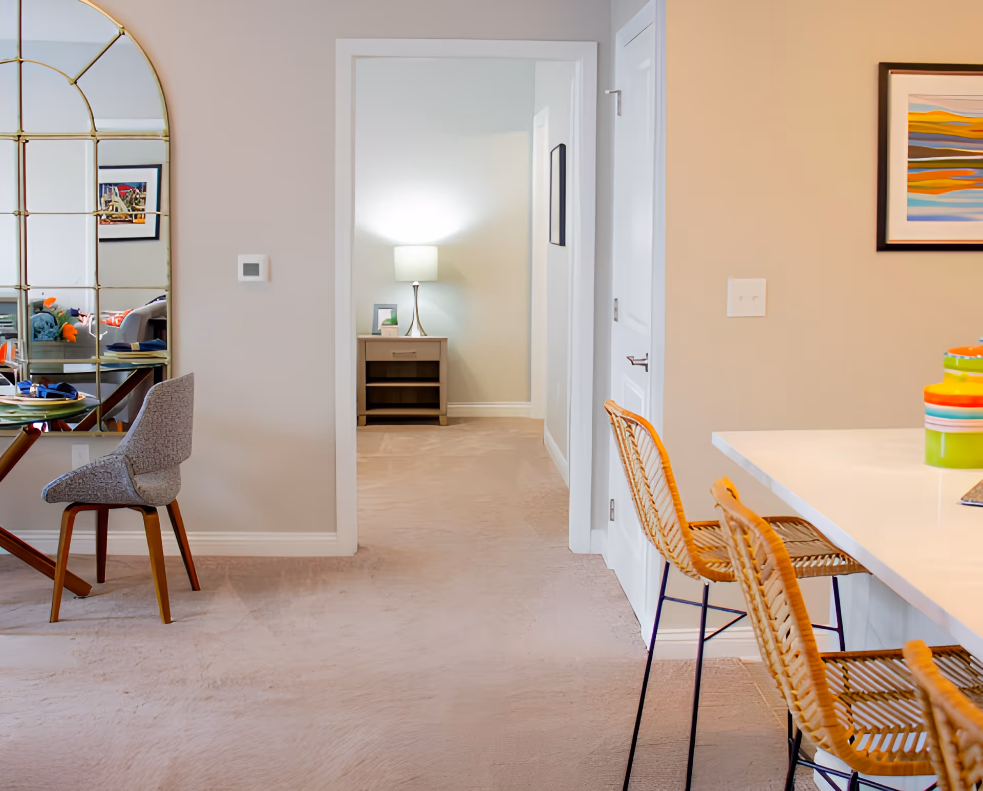 Open-plan interior with a kitchen counter and bar stools, a grey chair, and a hallway leading to a lamp-lit bedside table.
