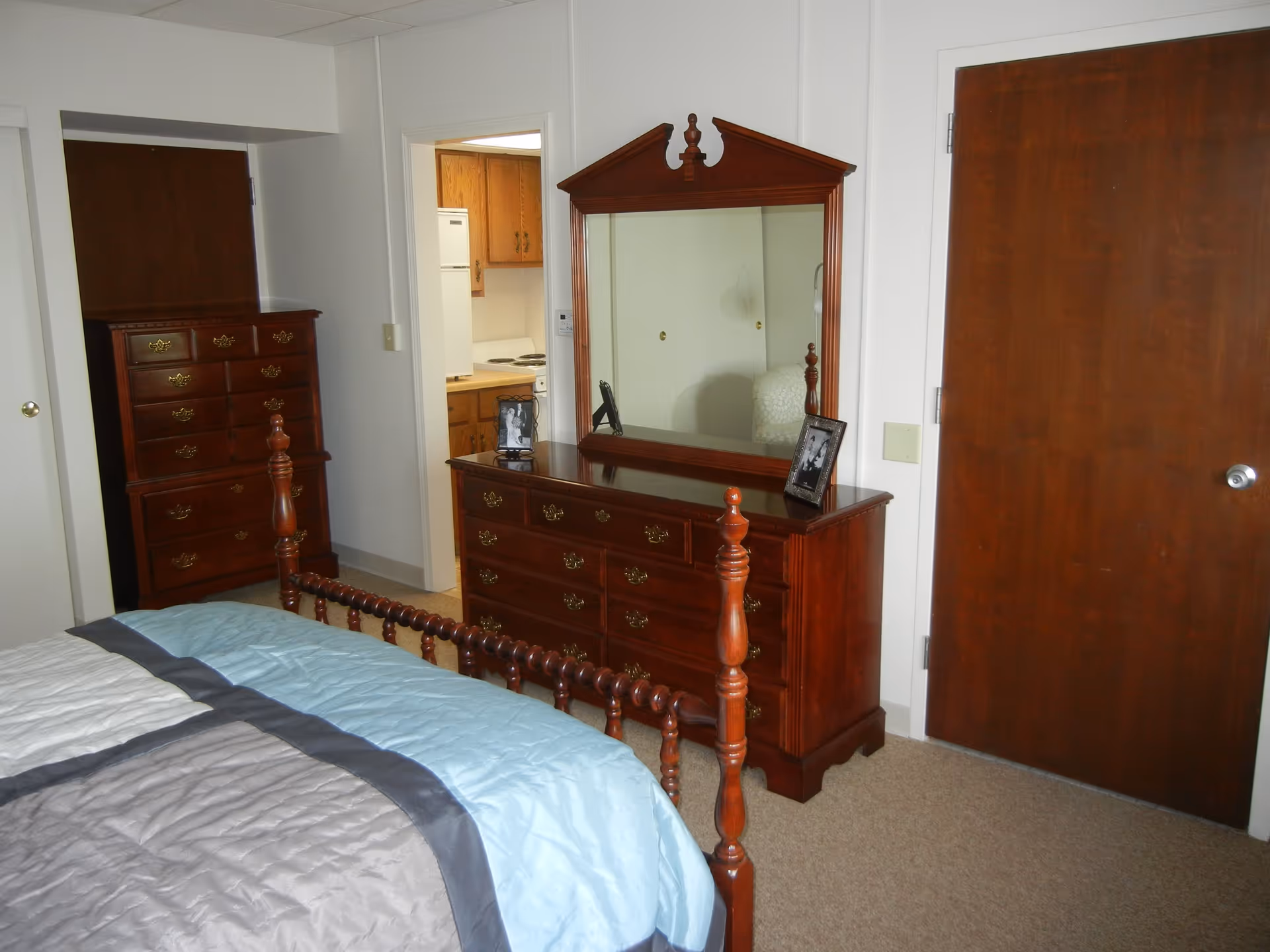 A bedroom with a wooden bed frame partially visible in the foreground, a wooden dresser with a large mirror and two framed photos on top, a tall wooden chest of drawers, and a doorway leading to a kitchen area with wooden cabinets and a white refrigerator. The walls are white and the floor is carpeted.