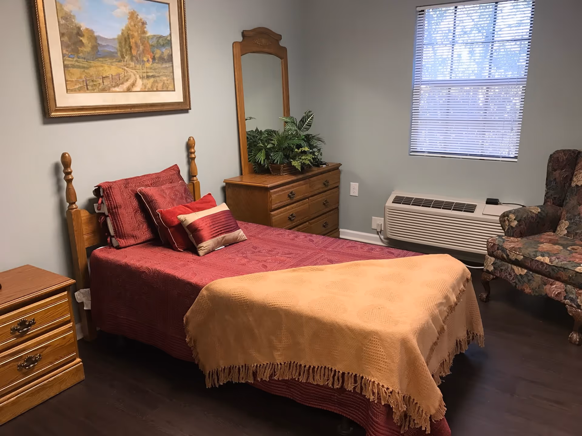 A cozy bedroom with a single wooden bed covered in a red bedspread and multiple pillows. There is a wooden dresser with a mirror and a plant on top, a wooden nightstand, a window with blinds, a floral upholstered armchair, and a framed landscape painting on the wall.