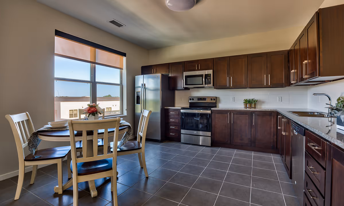 A modern kitchen with dark wooden cabinets, stainless steel appliances including a refrigerator, oven, microwave, and dishwasher. There is a round dining table with four chairs near a large window with a beige roller shade, and a small flower arrangement on the table.
