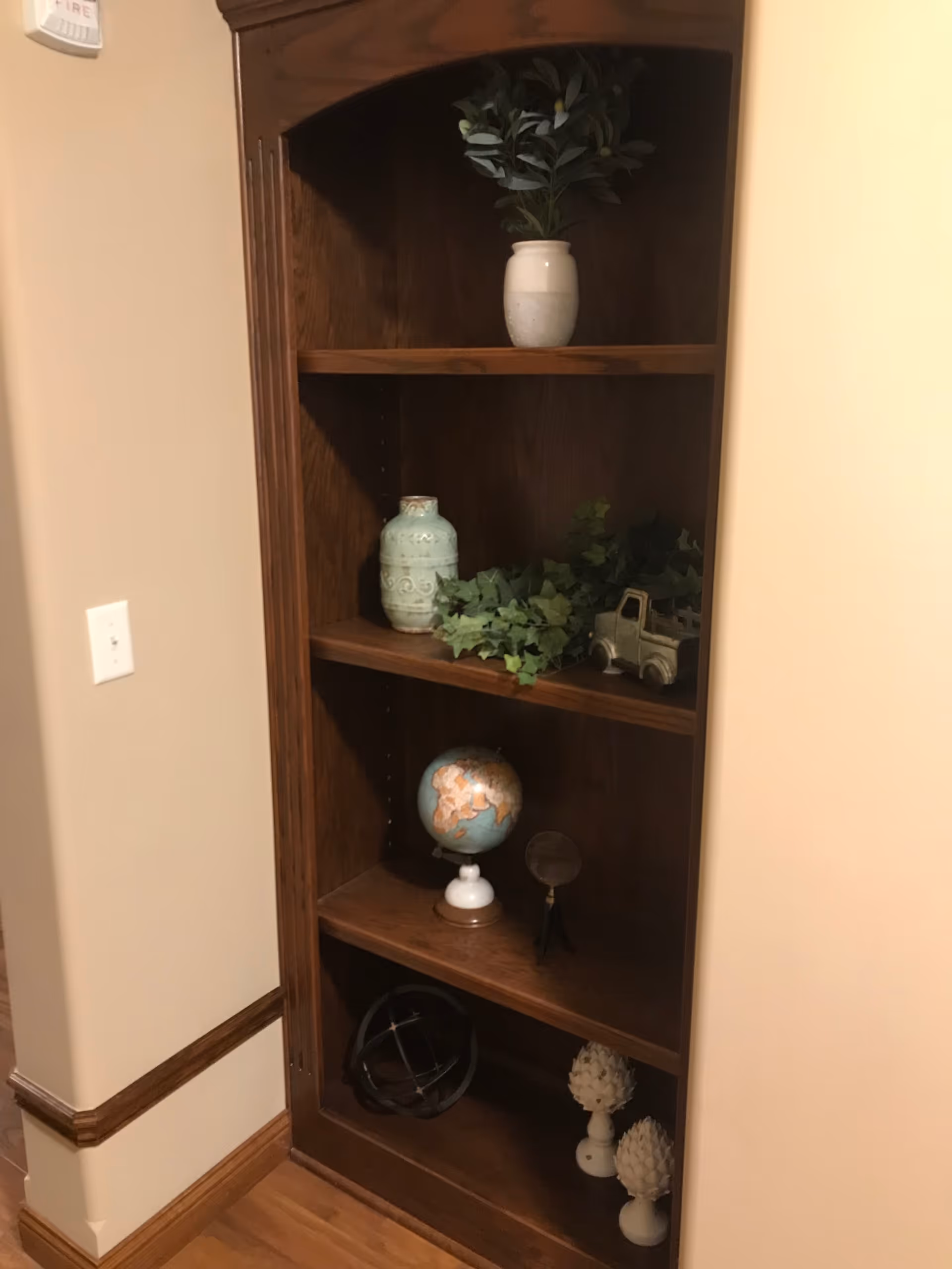 A wooden corner bookshelf with four shelves containing decorative items including a white vase with green leaves, a light blue ceramic jar, green ivy, a small toy truck, a globe, a magnifying glass on a stand, a black spherical armillary, and two white artichoke-shaped ornaments. The bookshelf is placed against beige walls with wooden trim and a hardwood floor.