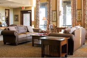 Communal living room with two sofas, a coffee table, side tables, lamps, and large windows with drapes.