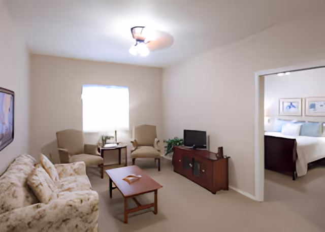 A cozy living room with a floral patterned sofa, two beige armchairs, a wooden coffee table, and a wooden TV stand with a small television. There is a window with a white curtain letting in natural light, a framed picture on the wall, and a doorway leading to a bedroom with a bed and framed artwork above it.