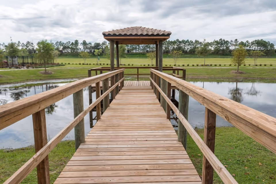 A wooden boardwalk with railings leading to a small covered gazebo over a pond, surrounded by grass and trees under a cloudy sky.