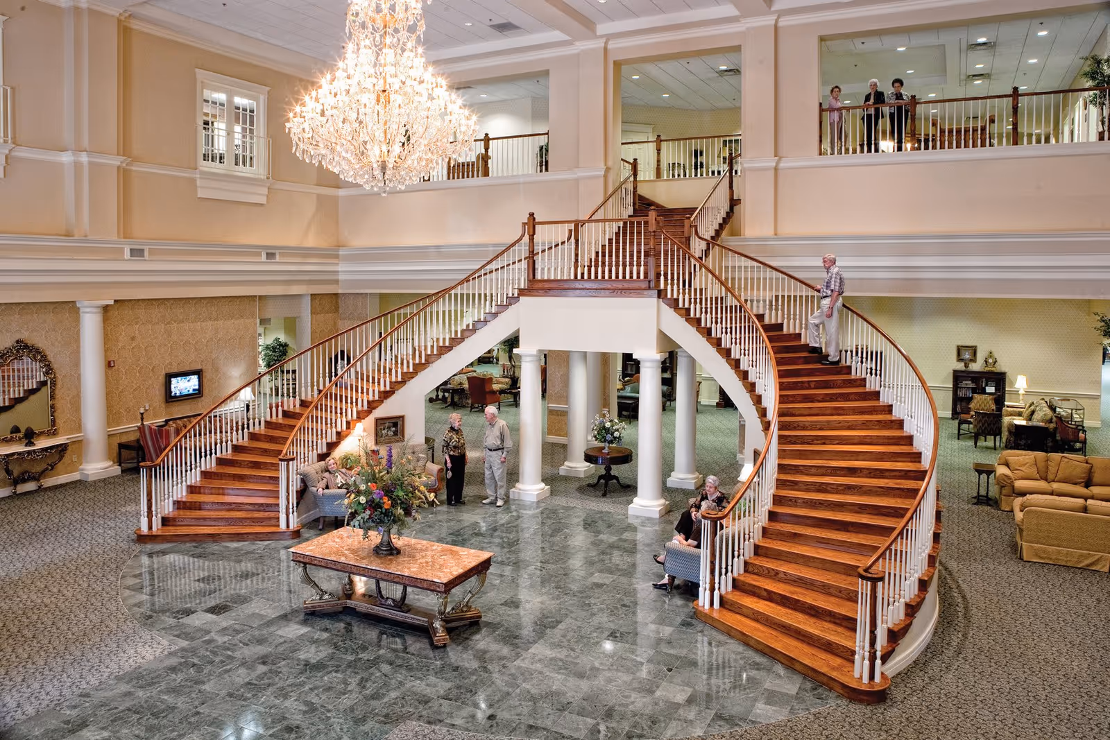 Spacious and elegant senior living community lobby with a grand double staircase featuring wooden steps and white railings. A large chandelier hangs from the ceiling, illuminating the area. Several seniors are seen sitting and standing, engaging in conversation. The floor is a mix of green marble and carpet, with a central table adorned with a floral arrangement. The upper level has a balcony overlooking the lobby.