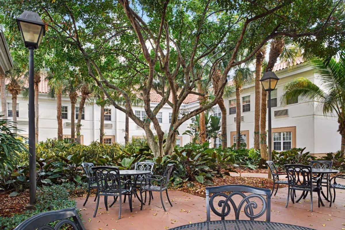 Outdoor courtyard area with black metal tables and chairs surrounded by lush green plants and trees, with a white two-story building in the background and lampposts providing lighting.