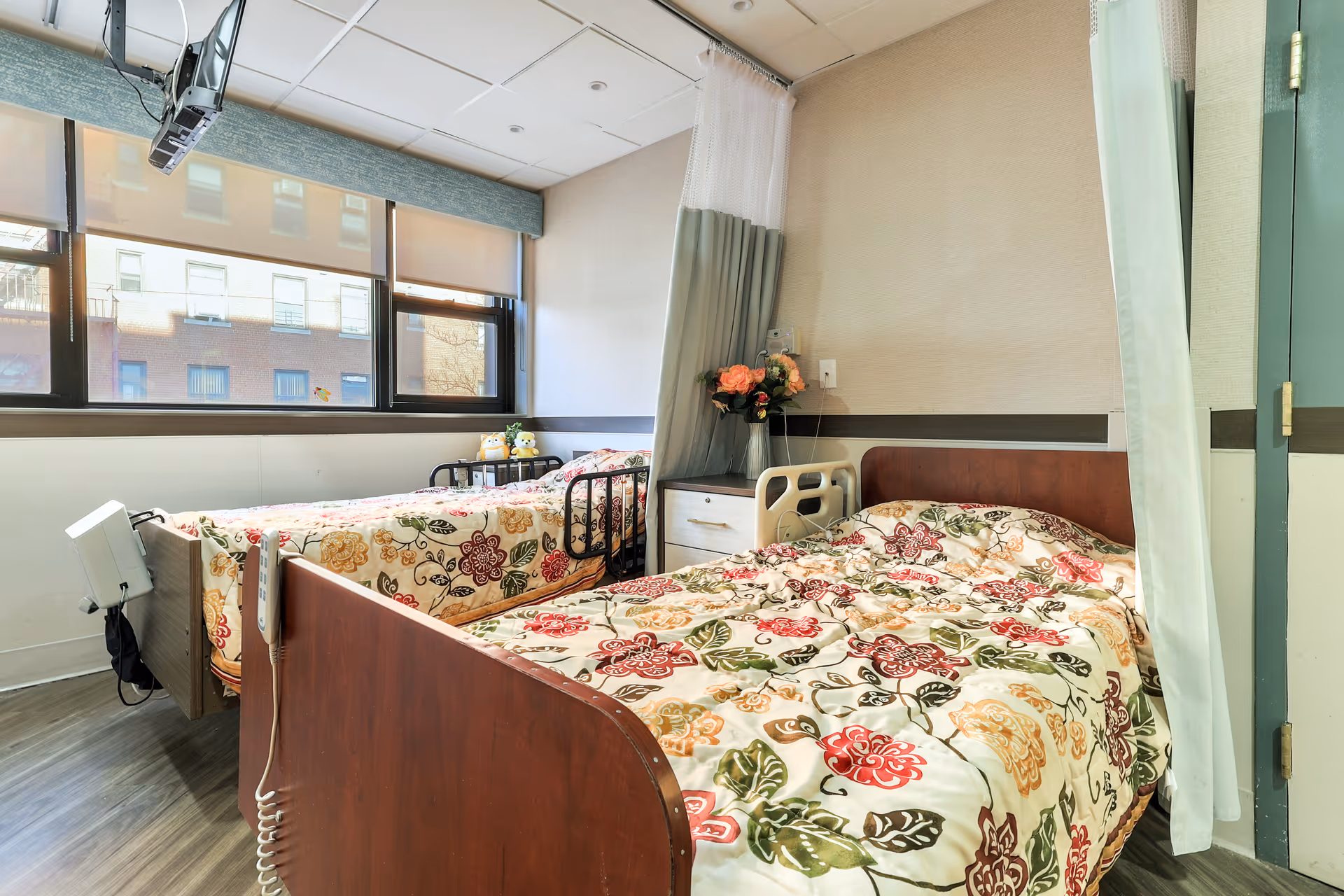 A bright room with two hospital-style beds featuring floral patterned bedspreads. There is a nightstand with a vase of flowers between the beds, a privacy curtain partially drawn, and large windows letting in natural light. The room has a clean and simple design with wood flooring and light-colored walls.