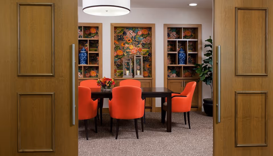 Wood-paneled double doors open to a dining area with a dark wood table surrounded by orange upholstered chairs and decorative shelving with colorful wallpaper.