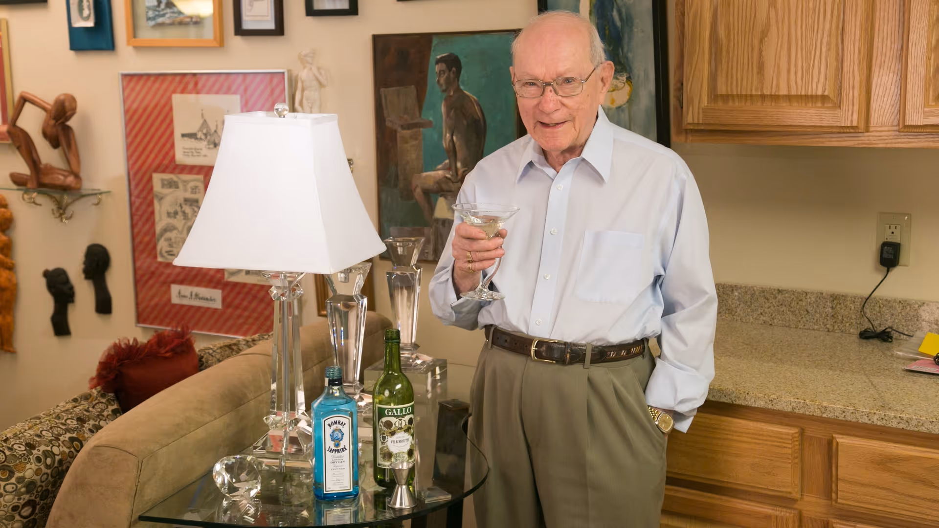 An elderly man wearing glasses, a light blue button-up shirt, and khaki pants stands in a cozy living area holding a cocktail glass. Behind him is a kitchen counter with wooden cabinets, and to his side is a glass table with a white lamp, a bottle of Bombay Sapphire gin, a bottle of Gallo wine, and a jigger. The wall behind features various framed artworks and sculptures.