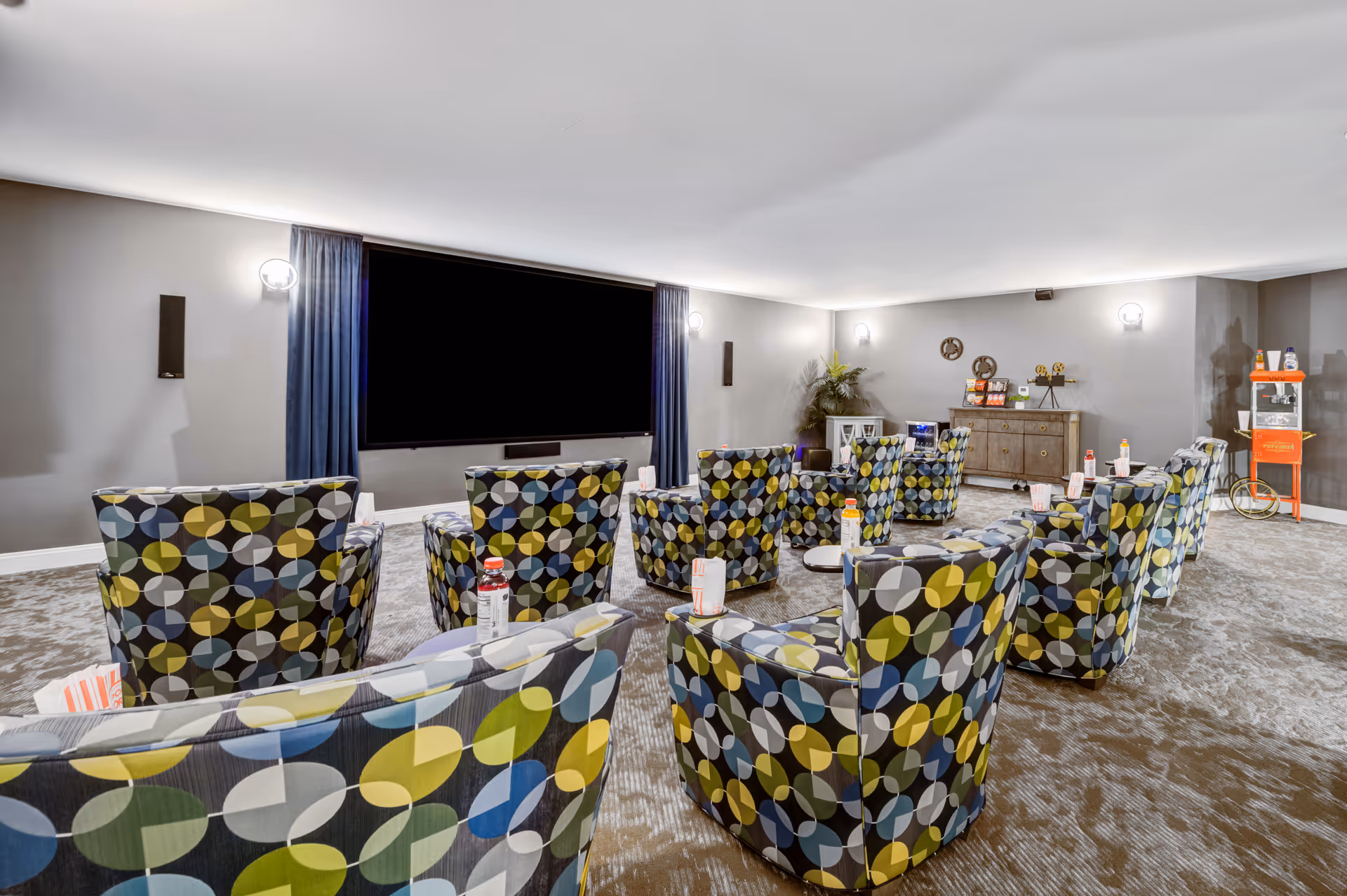 A cozy screening room with patterned swivel chairs arranged toward a large blank projection screen, with a popcorn machine and snack area at the back.