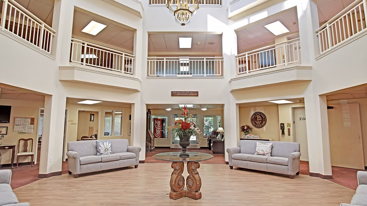 A spacious and well-lit common area in a senior living facility with two gray sofas facing each other, a round glass-top table with a decorative floral arrangement in the center, and a high ceiling with white railings on the upper level. The room has light-colored walls and wood flooring, with multiple doorways leading to other rooms. A Coca-Cola vending machine is visible in the background.