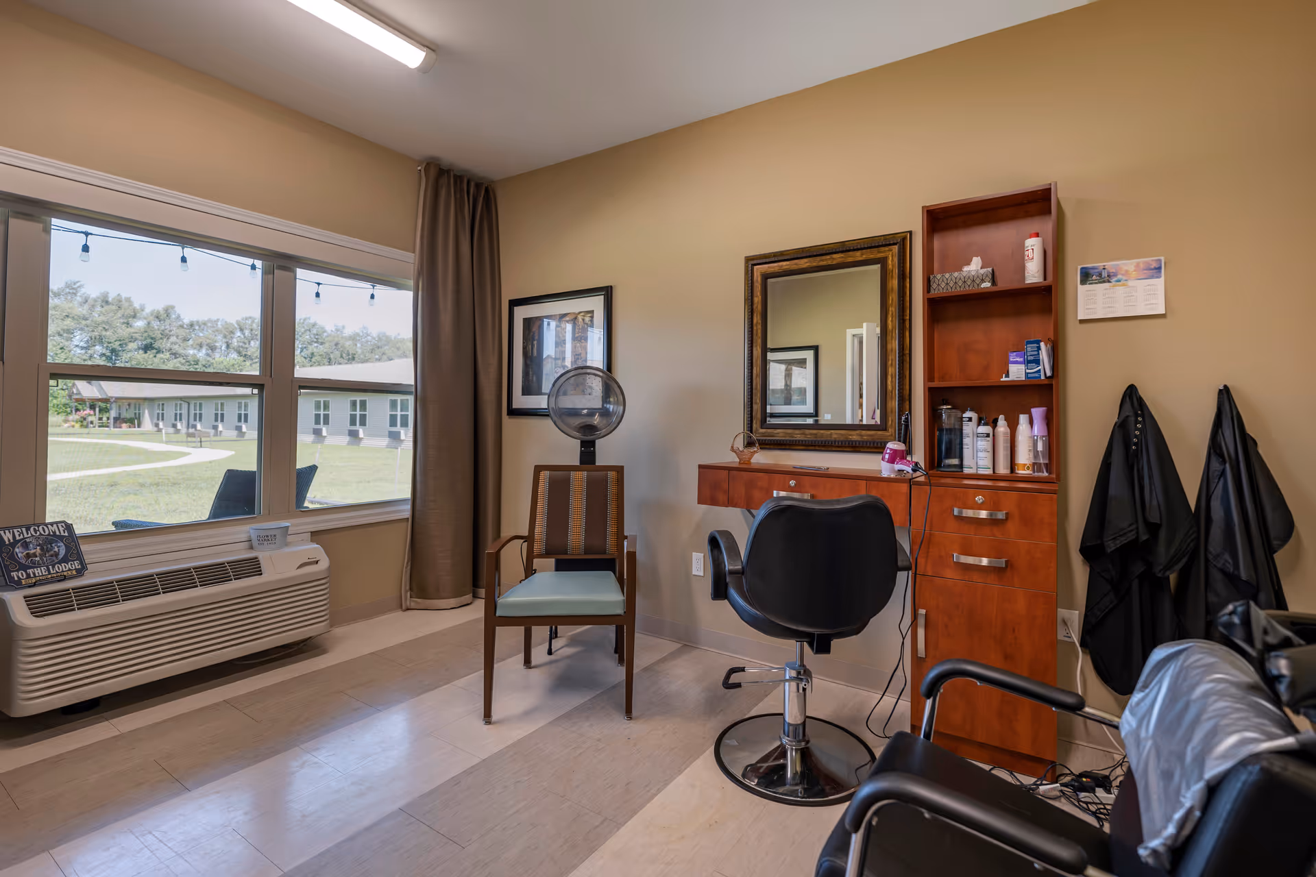 Interior of a salon area in a senior living facility with a large window showing an outdoor view. The room has a hair dryer chair, a styling chair in front of a mirror, a wooden cabinet with hair products, and black salon capes hanging on the wall.