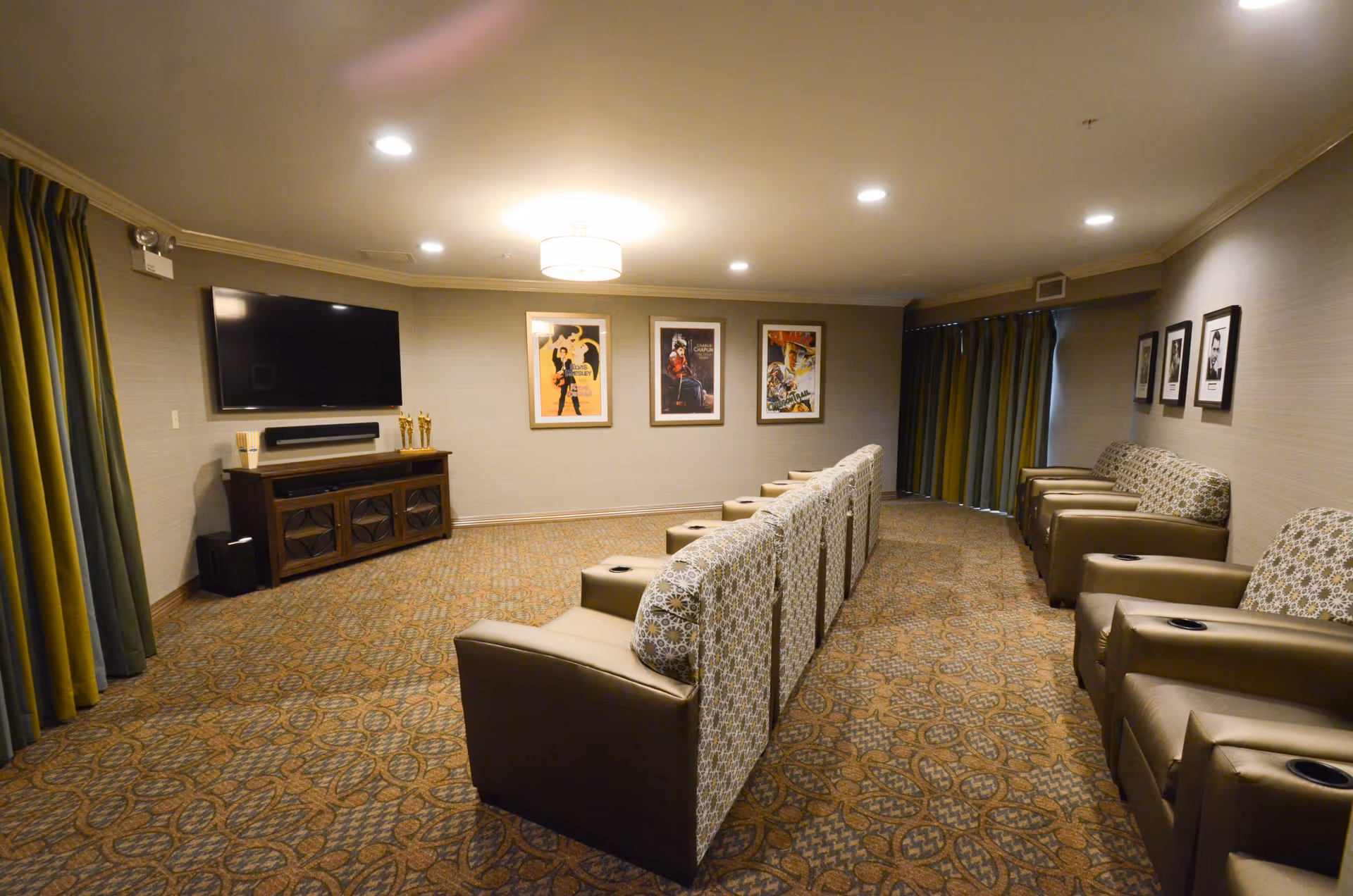 A cozy media room with two rows of cushioned armchairs facing a large flat-screen TV mounted on the wall. The room has patterned carpet, framed movie posters on the walls, and curtains covering the windows.