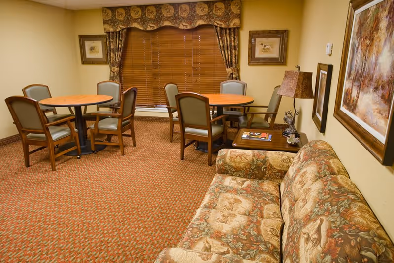 A cozy senior living common room with a patterned sofa, small round tables and chairs, framed artwork and a lamp.