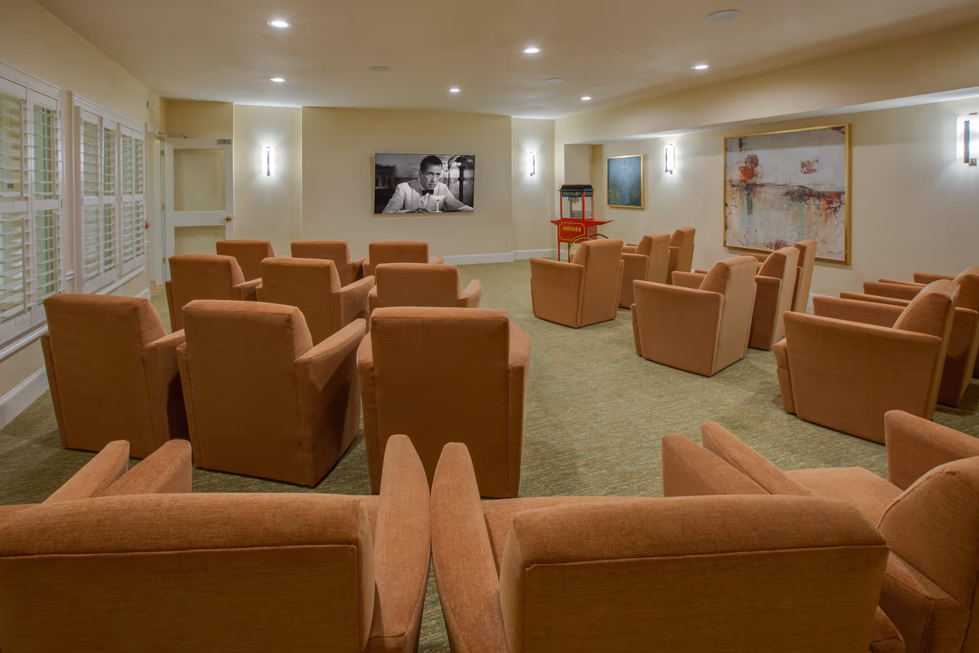 A small theater room with multiple rows of orange upholstered armchairs facing a wall-mounted TV screen showing a black and white image of a man. The room has beige walls, carpeted floor, wall sconces for lighting, a popcorn machine in the corner, and two framed artworks on the walls.