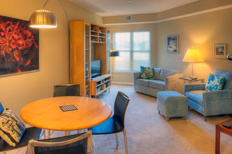 A cozy living room with a round wooden table surrounded by four chairs, a blue armchair with a matching ottoman, a small sofa, a wooden entertainment center with a TV, and a floor lamp. The room has beige walls, a window with blinds, and framed artwork on the walls.