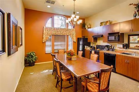 A dining room with a long table, chairs, and a kitchen area in the background.