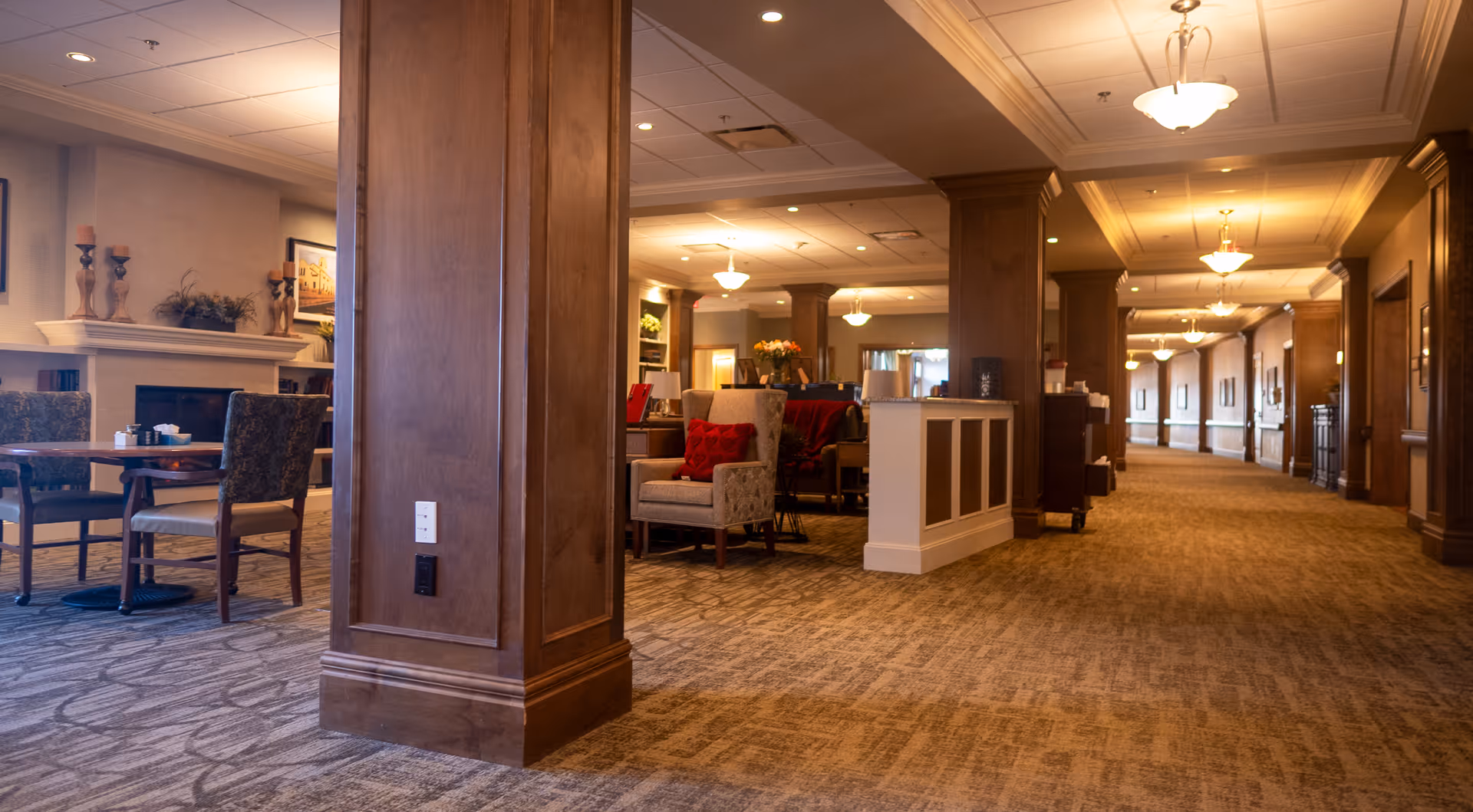 Interior view of a senior living facility common area with carpeted floors, wooden pillars, comfortable chairs with red cushions, tables, and a fireplace with decorative items on the mantel. The space is warmly lit with ceiling lights and has a long hallway extending into the background.