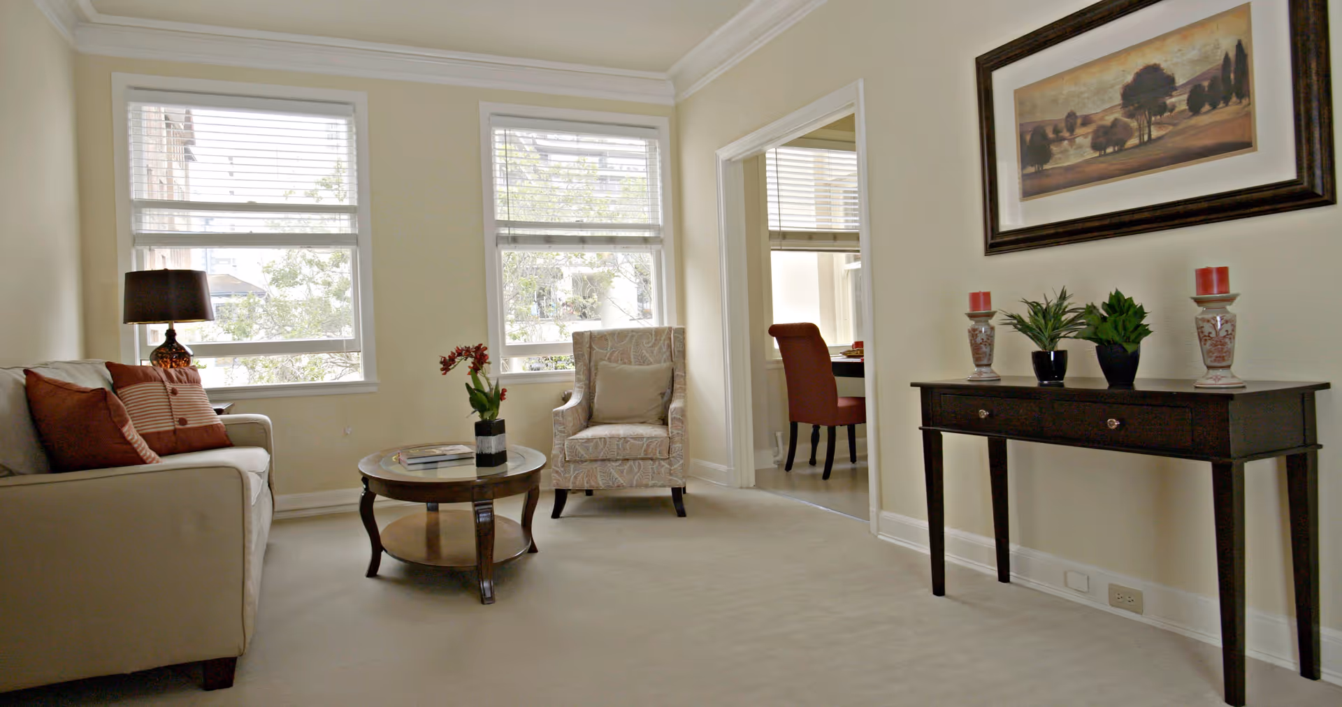 A bright living room with two large windows featuring white blinds. The room contains a beige sofa with red and striped pillows, a round wooden coffee table with a small plant and books, and a patterned armchair with a beige cushion. A dark wooden console table against the wall holds two decorative candle holders with red candles and two small potted plants. Above the console table hangs a framed landscape painting. Through an open doorway, a red chair and part of a dining table are visible in an adjacent room.