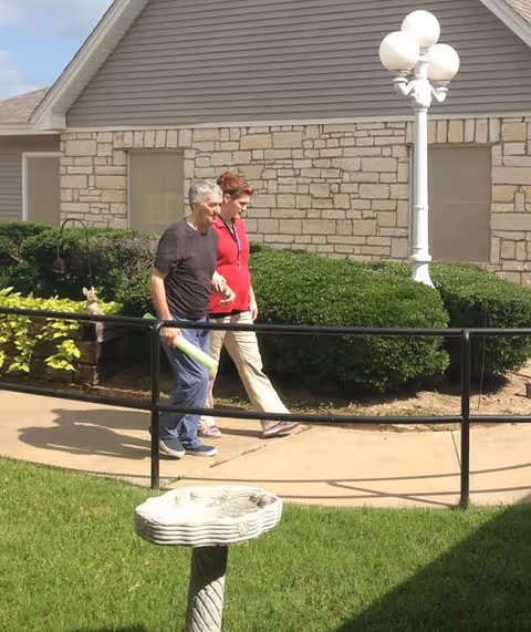 An elderly man and a woman walking together on a paved path outside near a building with stone and siding walls. There are green bushes, a white lamp post, and a birdbath in the grassy area nearby.