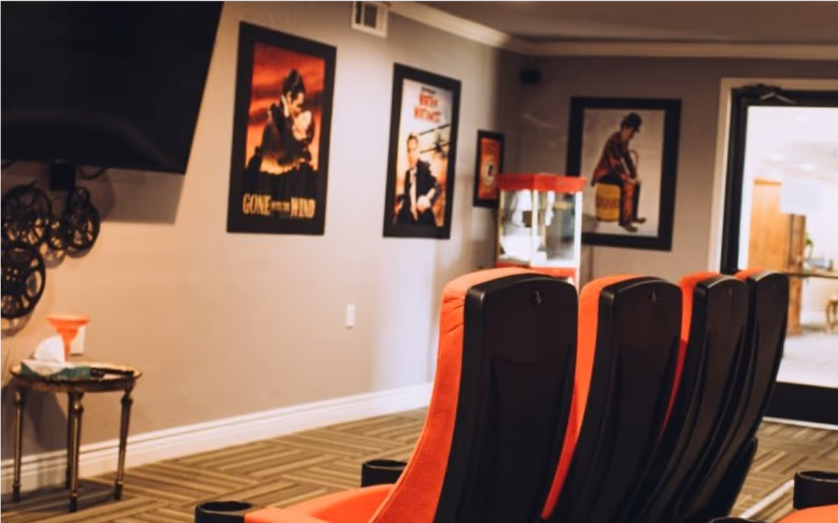 A small movie theater room with four red and black theater seats facing a wall-mounted TV. The walls are decorated with framed classic movie posters including Gone with the Wind and The Little Mermaid. There is a popcorn machine in the corner and a small side table with tissues and a funnel.