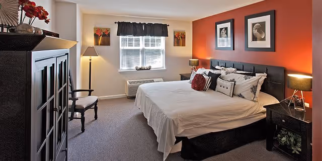 A well-lit bedroom with a large bed featuring white bedding and multiple pillows. The room has a red accent wall behind the bed with two framed pictures. There are two nightstands with lamps on either side of the bed, a window with blinds and a dark valance, a floor lamp, a wooden chair, and a dark wooden cabinet with decorative items on top.