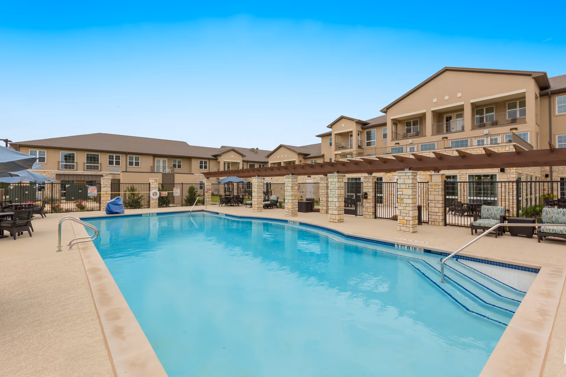 Outdoor swimming pool area at The Enclave at Round Rock Senior Living with clear blue water, surrounded by a beige concrete deck. There are several lounge chairs and tables with umbrellas around the pool. In the background, there is a multi-story senior living building with balconies and a pergola structure supported by stone columns.
