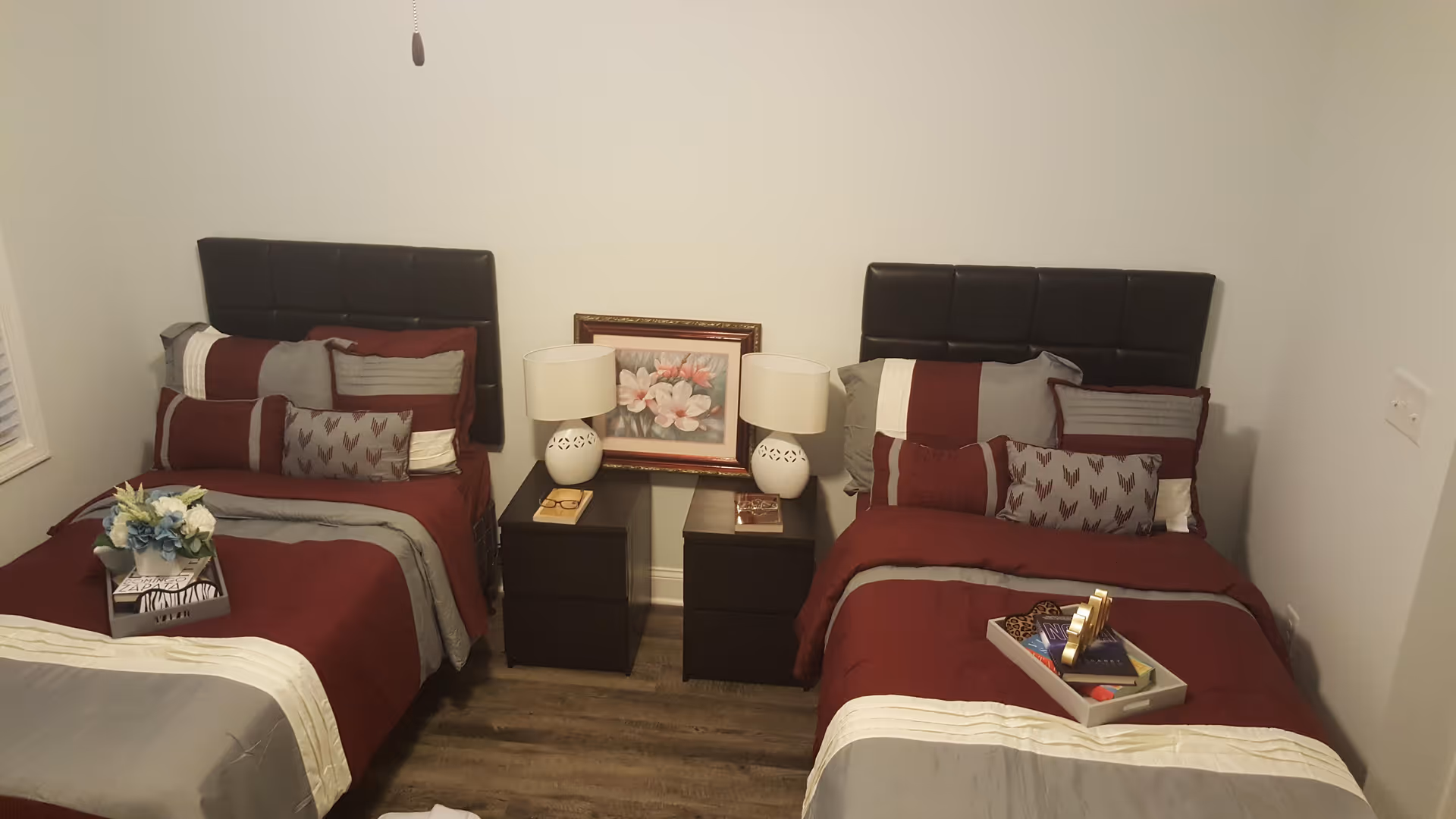 A bedroom with two single beds, each with a dark headboard and red, gray, and white bedding. Between the beds are two dark nightstands with white lamps and a framed floral picture on the wall. One bed has a tray with books and decorative items, and the other has a small flower arrangement on a box.