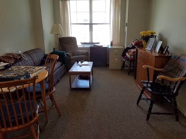 A cozy living room with a plaid sofa, a wooden coffee table, an armchair, and a wooden dining table with chairs. There is a window with blinds letting in natural light. A wooden dresser with framed photos and a vase of yellow flowers is on the right side, along with a rocking chair draped with a blanket. A filing cabinet and a small table with items are near the window.