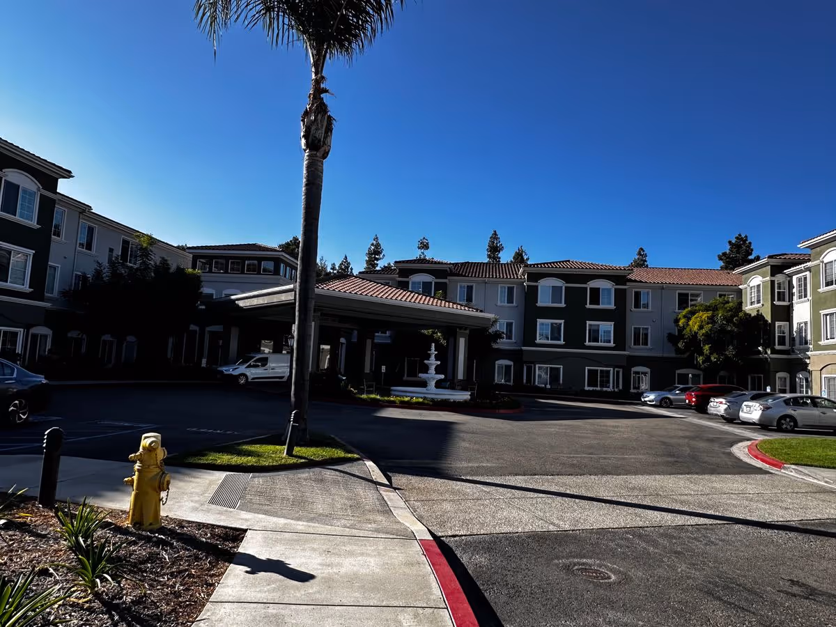 Front entrance of a three-story senior living building with a covered driveway, fountain, palm tree, and parked cars under a clear blue sky.