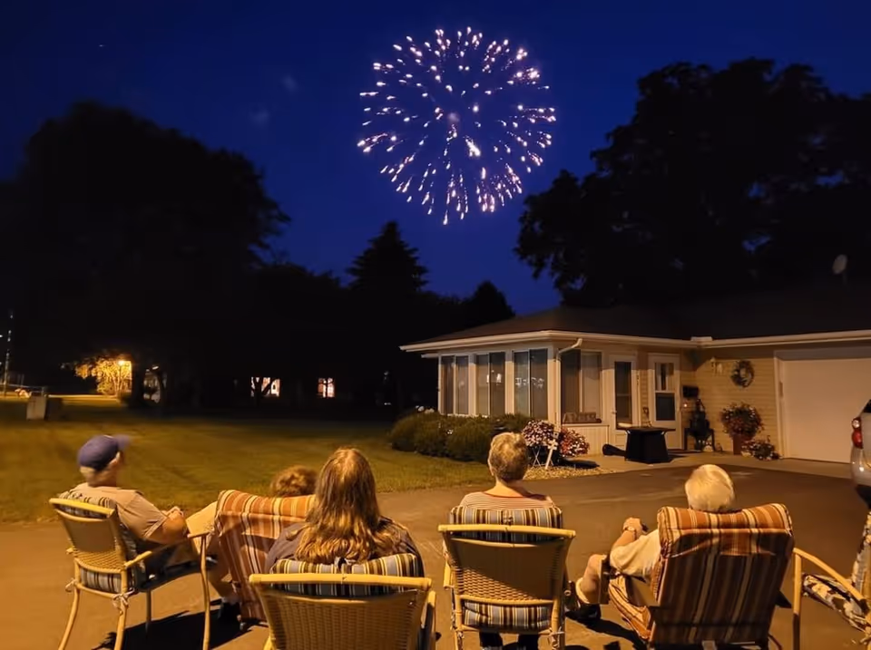 Five people sitting on outdoor chairs in a driveway at night watching fireworks in the sky above a single-story house with a well-lit yard and trees in the background.