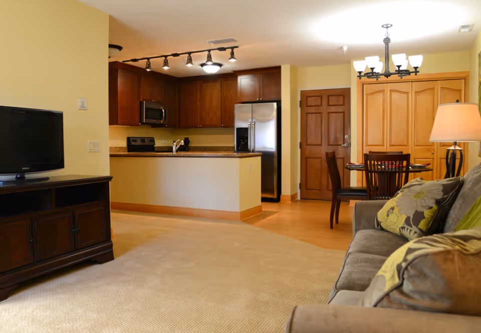 Interior view of a senior living community apartment showing a living room with a gray couch and floral pillows, a wooden TV stand with a flat-screen TV, a dining area with a wooden table and chairs, and a kitchen with wooden cabinets, a stainless steel refrigerator, stove, and microwave.