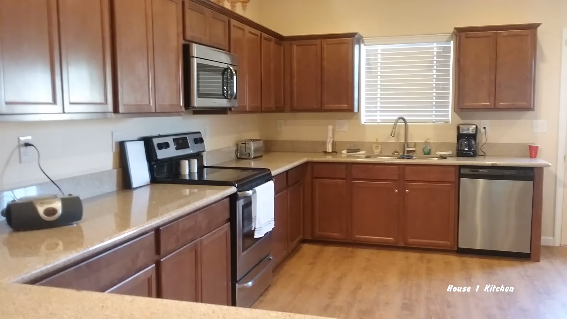 A kitchen with wooden cabinets, a stainless steel stove with a microwave above it, a toaster, a coffee maker, a dishwasher, and a window with blinds. The countertops are light-colored, and the floor is wooden.