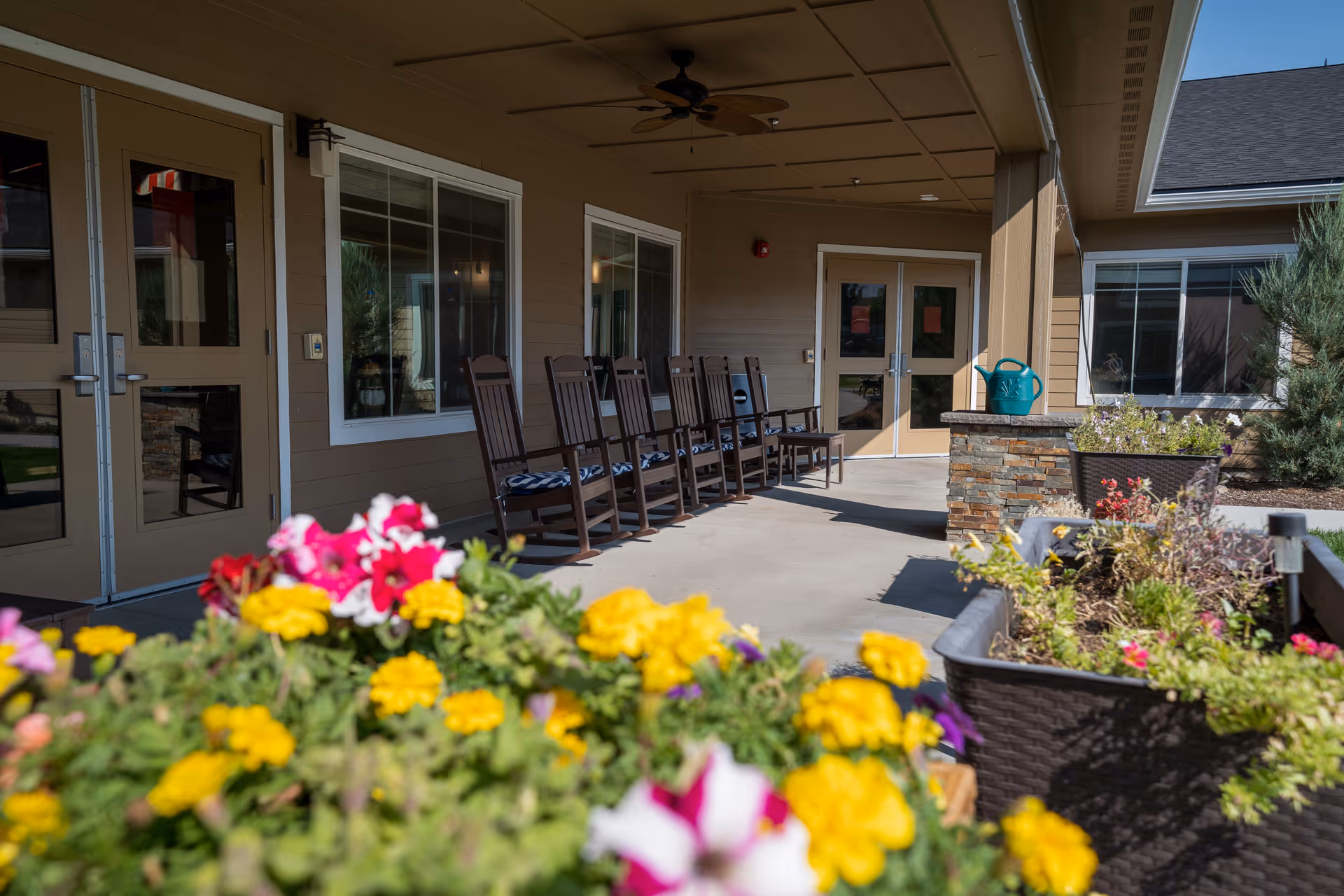 Outdoor covered patio area with a row of wooden rocking chairs along the wall of a building. In the foreground, there are colorful flowers in planters, and a watering can is visible on a stone ledge. The building has beige siding and multiple windows and doors.
