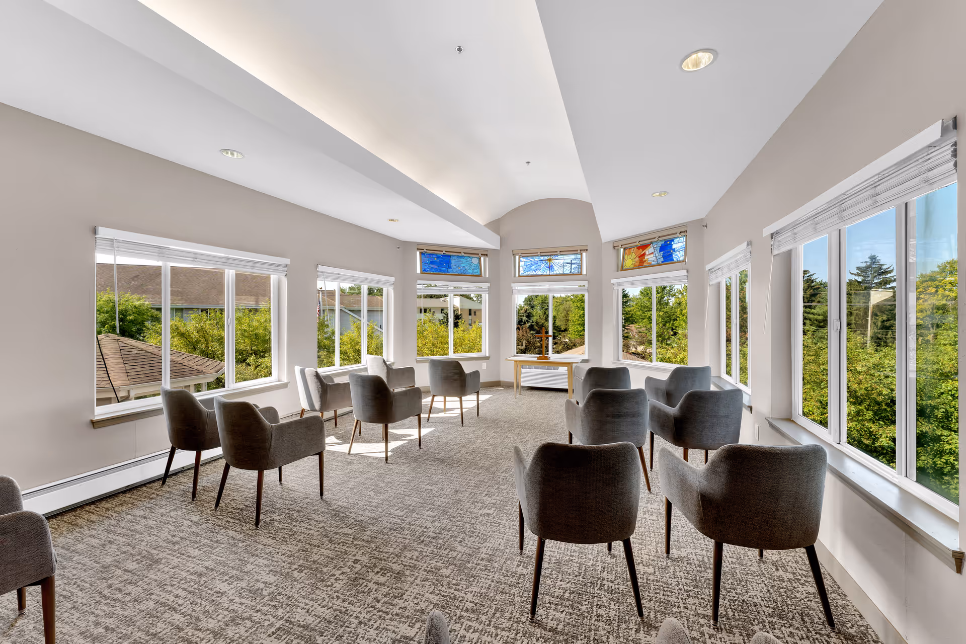 Bright lounge with rows of gray armchairs arranged facing large windows overlooking trees.