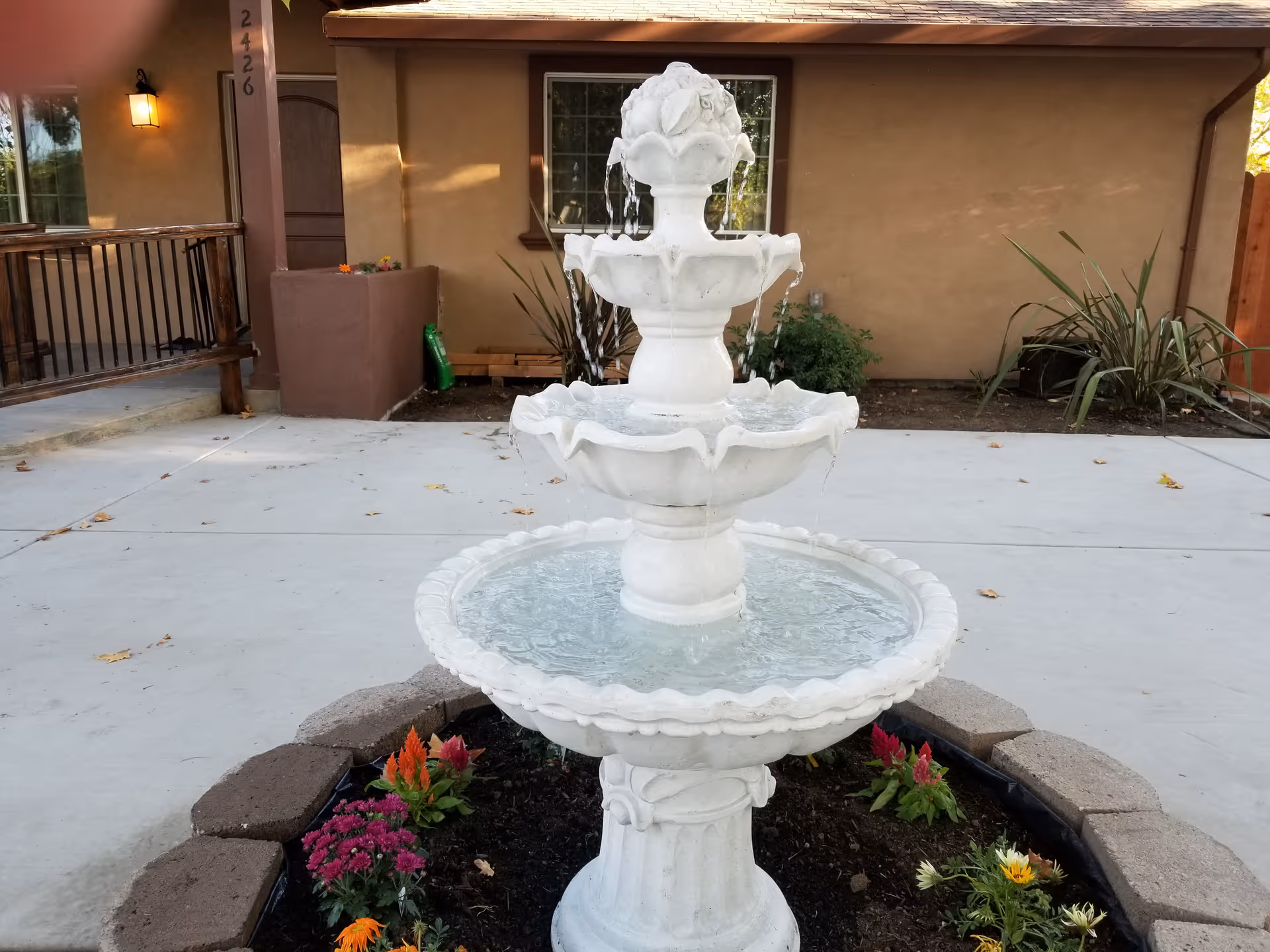 A white three-tiered water fountain surrounded by a circular flower bed with colorful flowers, situated on a concrete patio in front of a beige building with a window and a wooden railing on the left side.