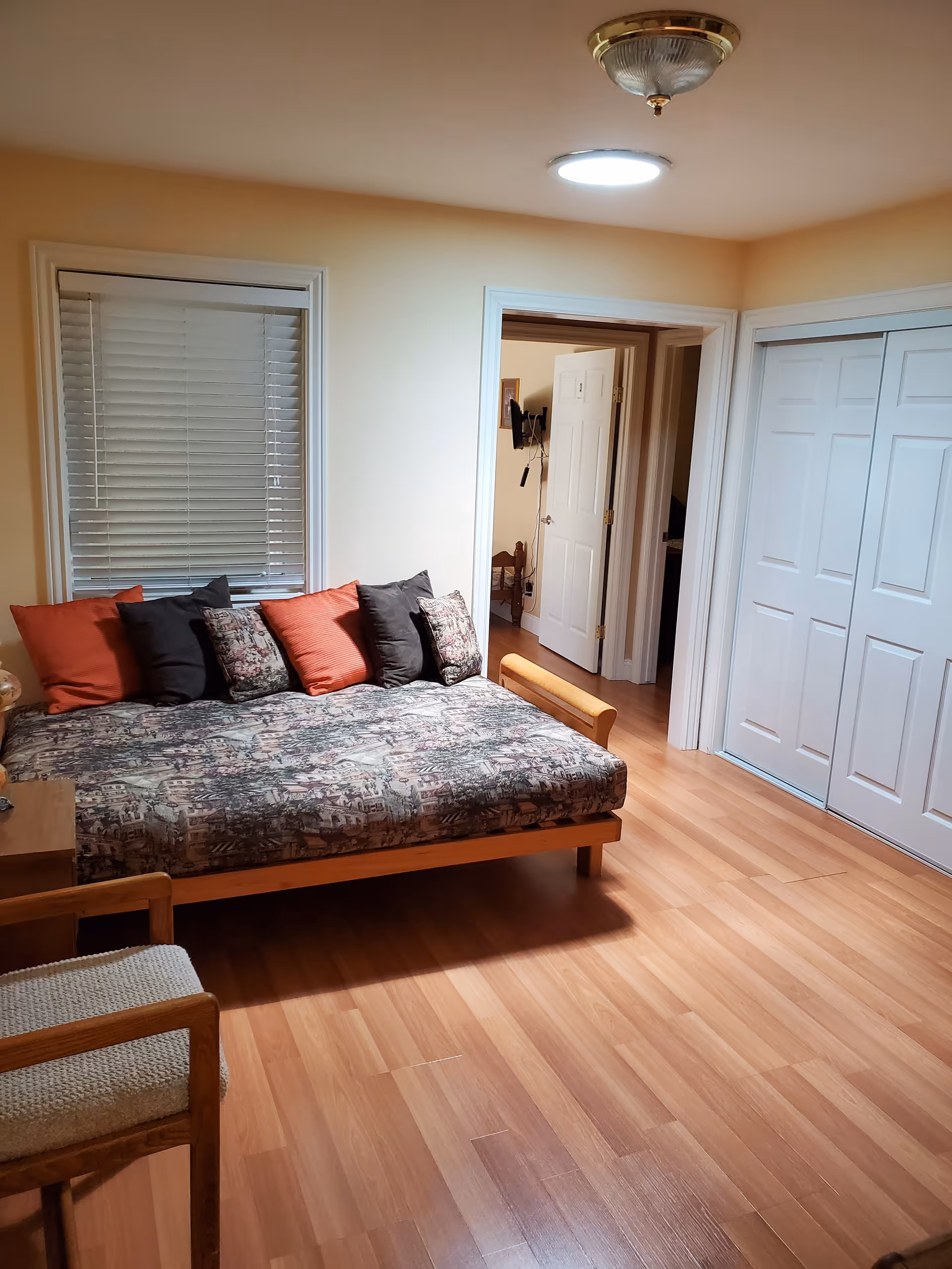 A cozy room with light wooden flooring, a daybed with patterned mattress and multiple cushions in black, orange, and floral designs. There is a wooden chair with a woven seat in the foreground. The room has a window with closed blinds, a ceiling light fixture, and white double closet doors. An open doorway leads to another room with a glimpse of a mounted TV and furniture.