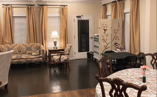 A cozy senior living common area with a patterned sofa and matching armchair near a side table with a lamp. There are large windows with beige curtains, a small table with chairs, and a round dining table with a floral tablecloth and a small flower vase. The room has dark flooring and light-colored walls.