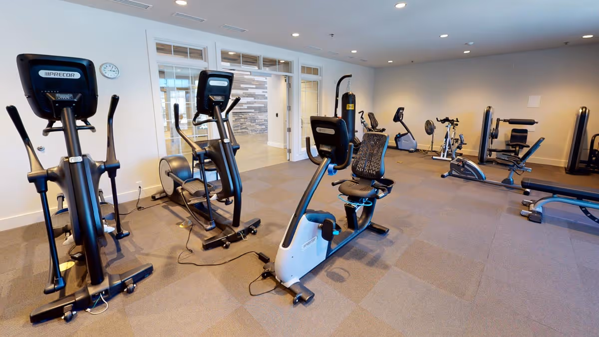 A fitness room with various exercise equipment including stationary bikes, elliptical machines, a punching bag, and weight machines. The room has carpeted flooring, white walls, and a clock on the wall. There is a glass door leading to another area with a stone accent wall visible through it.